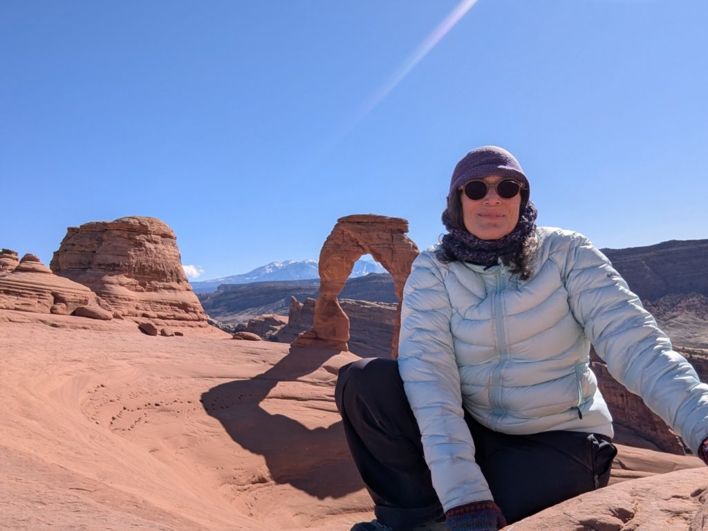 Woman bundled up against the cold in front of a stone arch,  and rocky landscape. Blue sky with a tiny cloud and a mountain range on the horizon.